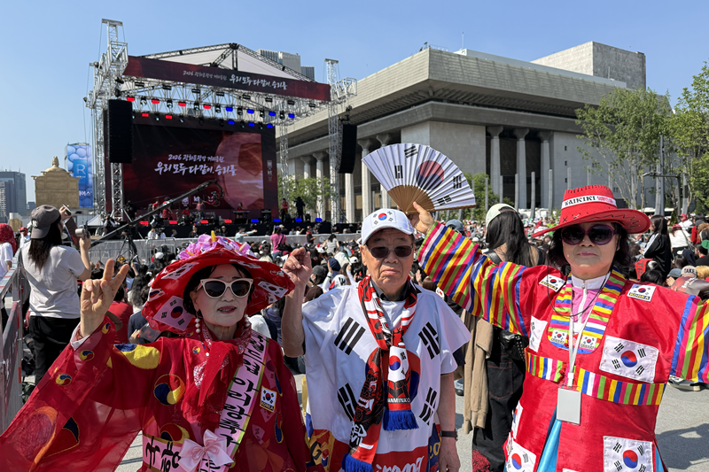 Trois supporters de l&rsquo;&eacute;quipe nationale cor&eacute;enne d&eacute;cor&eacute;s de drapeaux cor&eacute;ens, le taekeugki, sur la place Gwanghwamun, le 12 juin 2026. &copy; Hong Angie / Korea.net