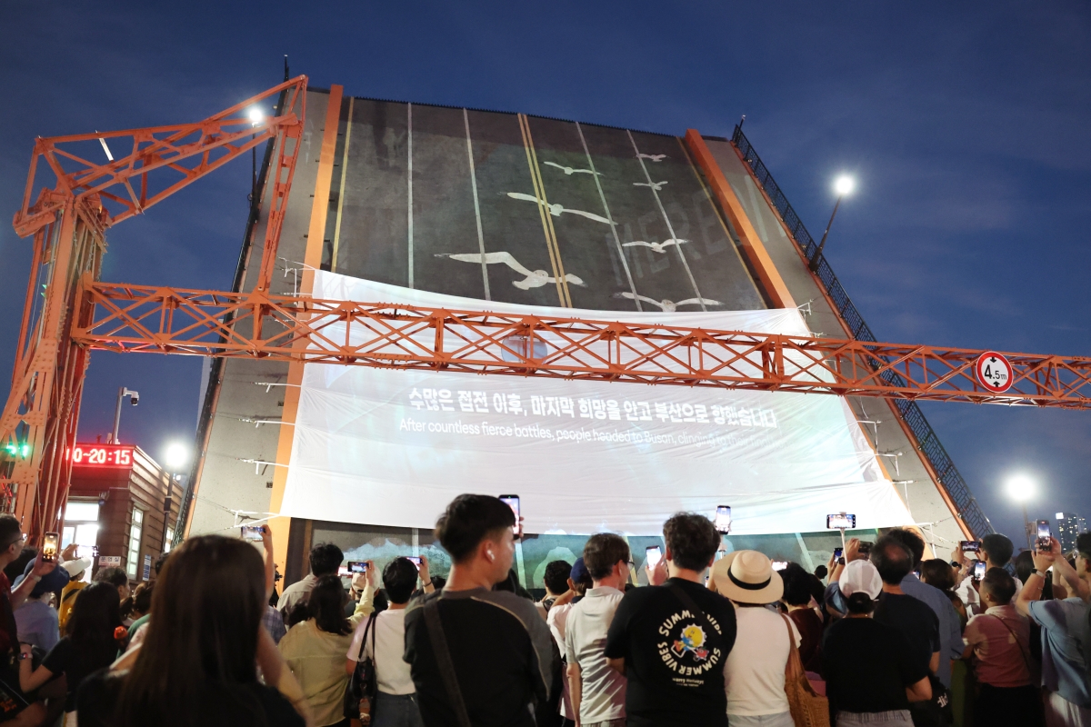 Un groupe de personnes assiste à une projection retraçant l'histoire de Busan pendant la guerre de Corée diffusée sur le pont Yeongdo en train d'être levé, lors du programme de visites guidées nocturnes Busan Heritage Night, le 25 juillet 2026. Construit en 1932, le pont Yeongdo fut le lieu de rassemblement des réfugiés de la guerre de Corée et de leurs familles affluant à Busan, alors capitale provisoire. © Park Dae-jin / Korea.net